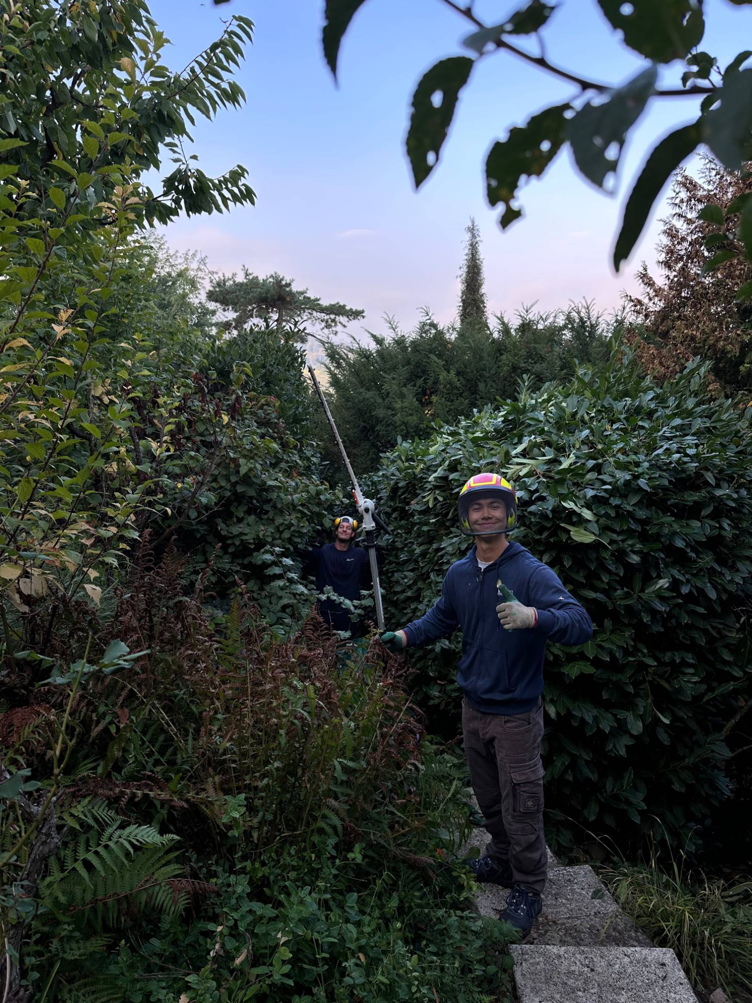 Zwei Teammitglieder mit Helm und Teleskop-Heckenschere zwischen hohen Hecken, eines zeigt den Daumen nach oben